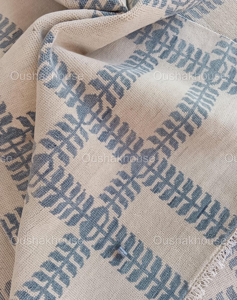 Op de afbeelding: Een geweven beige en blauw tapijt met een geometrisch patroon. Het tapijt heeft een franje rand.