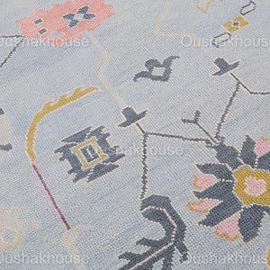 May include: Close-up of a light blue rug with a geometric pattern in shades of pink, yellow, and gray. The pattern features stylized flowers, leaves, and abstract shapes.