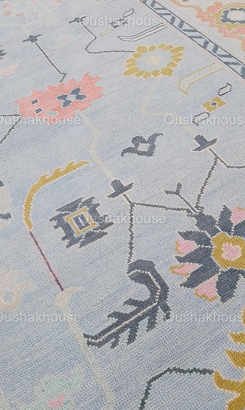 May include: Close-up of a light blue rug with a geometric pattern in shades of pink, yellow, and gray. The pattern features stylized flowers, leaves, and abstract shapes.