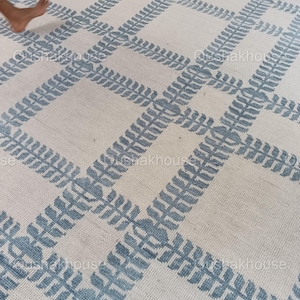 Op de afbeelding: Een wit tapijt met een blauw geometrisch patroon. Het patroon bestaat uit herhalende bladeren.