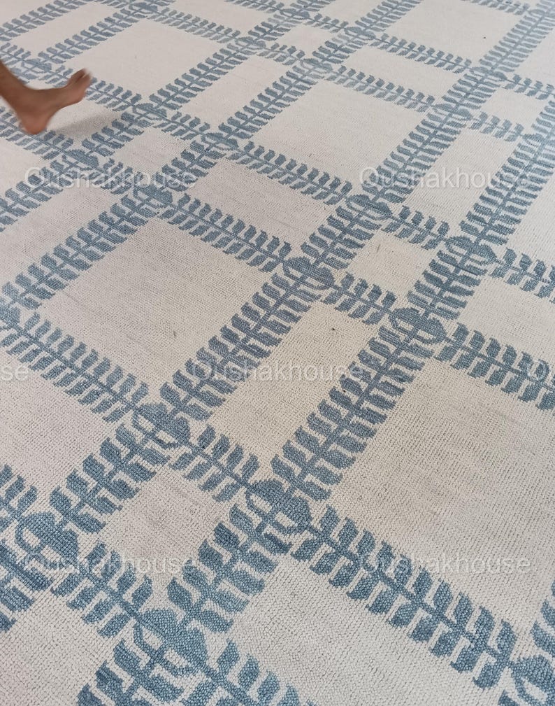 Op de afbeelding: Een wit tapijt met een blauw geometrisch patroon. Het patroon bestaat uit herhalende bladeren.