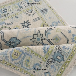 Pode incluir: Um tapete de cor creme com um padrão geométrico azul, verde e cinza. O tapete tem uma borda de franjas e está dobrado ao meio.