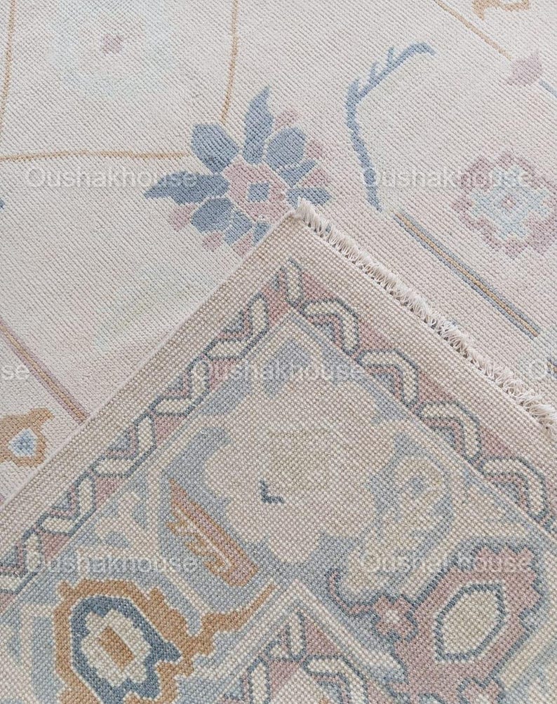 May include: Close-up of a cream-coloured rug with a detailed floral pattern in shades of blue, pink, and brown. The rug has a fringed edge.