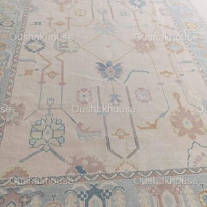 May include: A cream-coloured rug with a traditional Persian design featuring intricate floral patterns in shades of blue, pink, and beige. The rug has a border with a repeating geometric pattern.