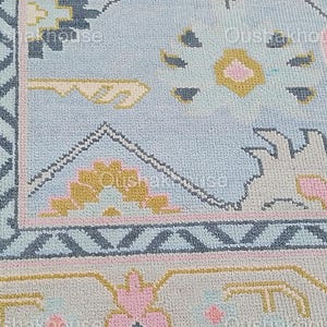 May include: A close-up of a light blue rug with a geometric pattern in shades of pink, yellow, and blue. The rug has a border with a zig-zag pattern.