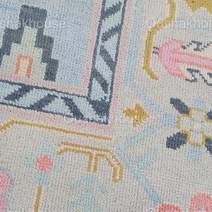 May include: A close-up of a light blue rug with a geometric pattern. The rug features pink, blue, and yellow accents. The pattern includes a house, flowers, and stars.