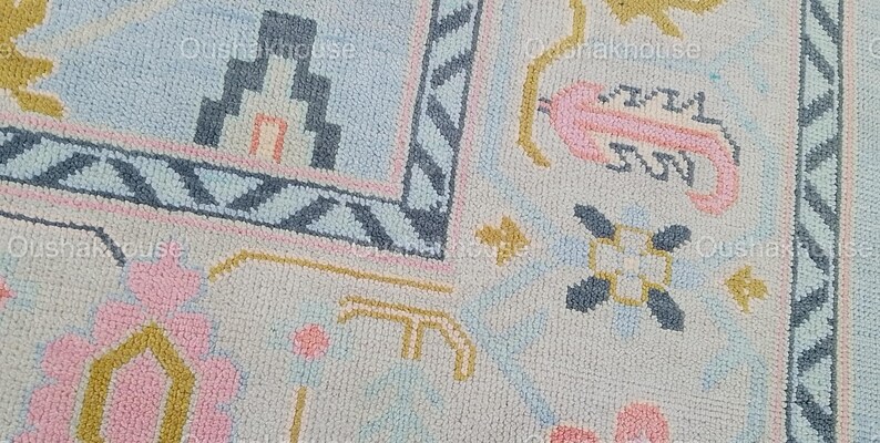 May include: A close-up of a light blue rug with a geometric pattern. The rug features pink, blue, and yellow accents. The pattern includes a house, flowers, and stars.