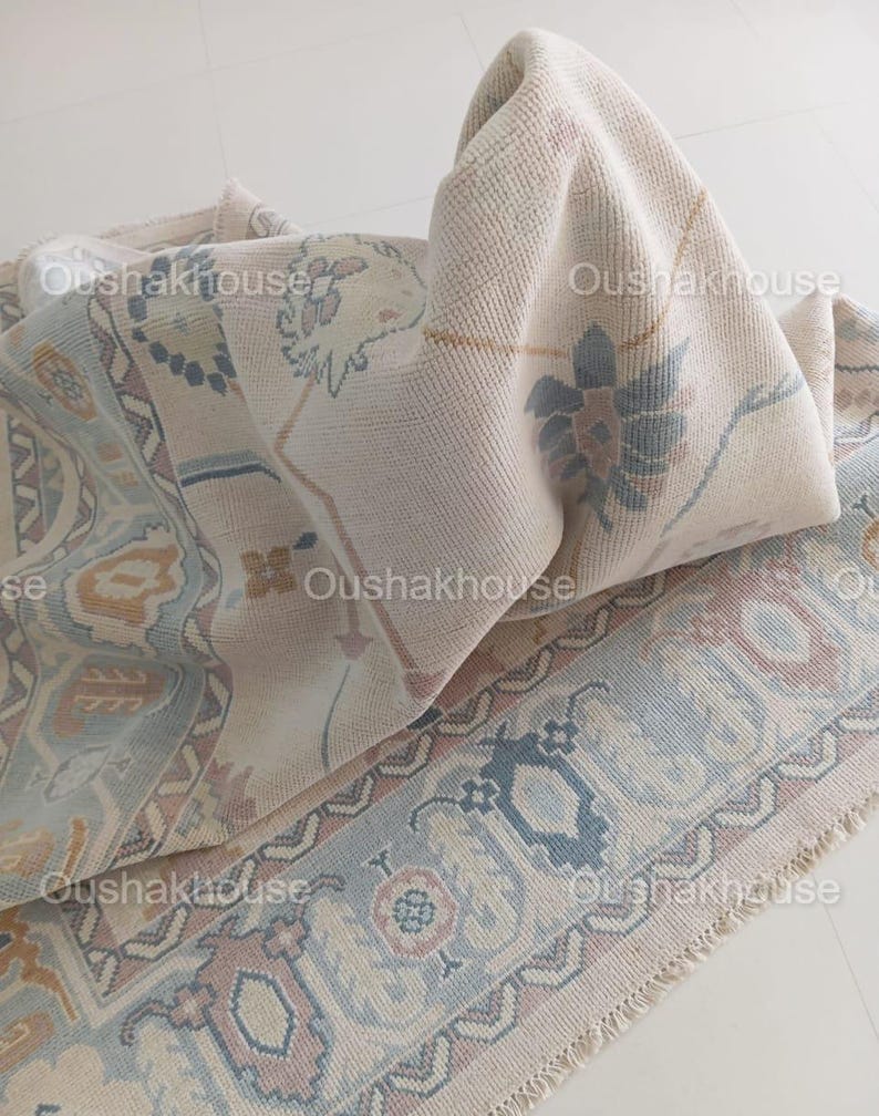 May include: A cream-coloured rug with a blue and pink floral pattern. The rug has a fringe border and is folded in half.