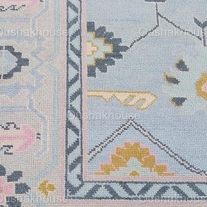 May include: Close-up of a light blue rug with a geometric pattern in shades of pink, yellow, and gray. The rug has a border with a zig-zag design.