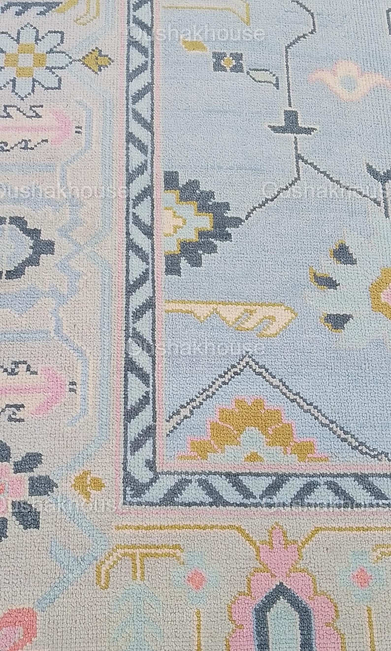 May include: Close-up of a light blue rug with a geometric pattern in shades of pink, yellow, and gray. The rug has a border with a zig-zag design.