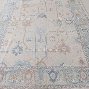 May include: A cream-coloured rug with a traditional Persian design in shades of blue, pink, and gold. The rug has a fringed border.