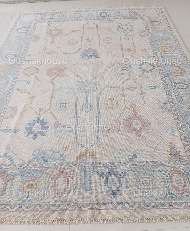 May include: A cream-coloured rug with a traditional Persian design in shades of blue, pink, and gold. The rug has a fringed border.