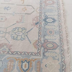 May include: A close-up of a cream-coloured rug with a detailed floral pattern in shades of pink, blue, and yellow. The rug has a fringed edge and a subtle distressed look.