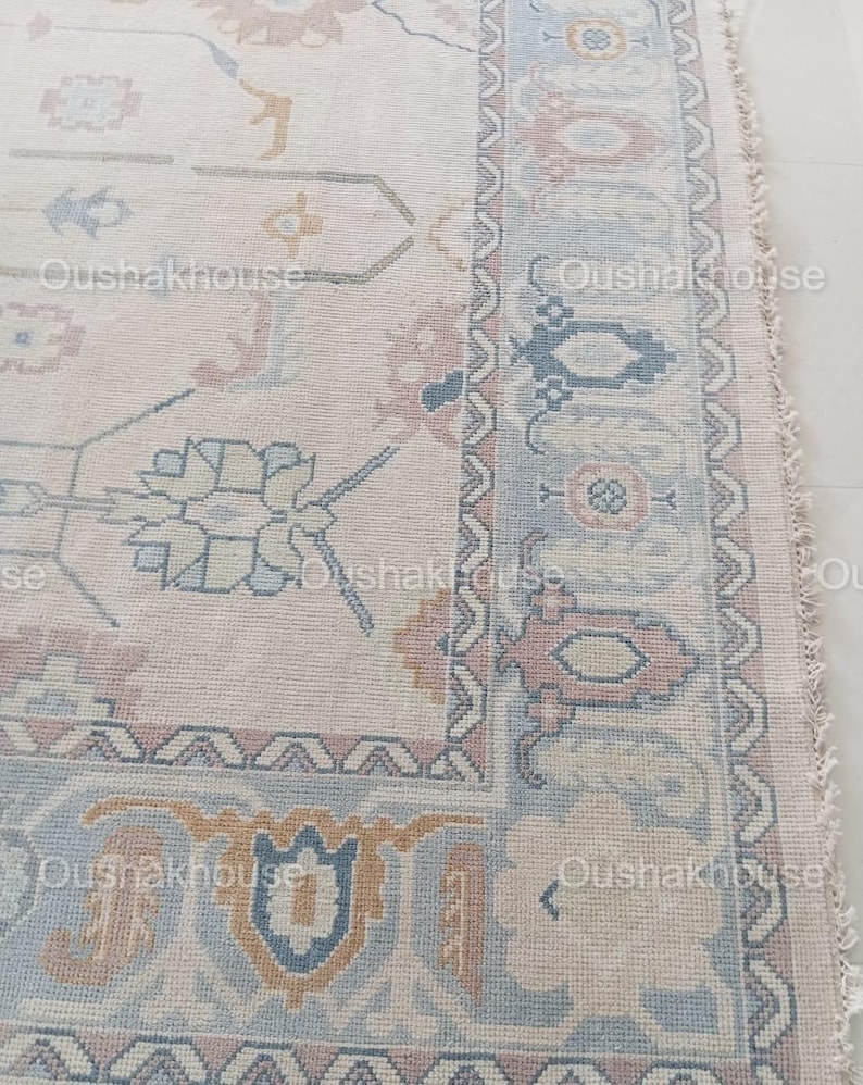 May include: A close-up of a cream-coloured rug with a detailed floral pattern in shades of pink, blue, and yellow. The rug has a fringed edge and a subtle distressed look.