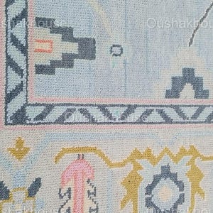 May include: A close-up of a light blue rug with a geometric pattern in shades of pink, yellow, and gray. The rug has a fringe border.