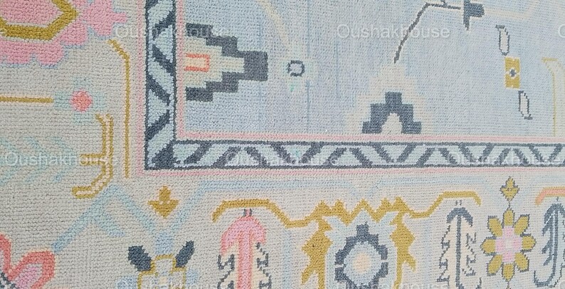 May include: A close-up of a light blue rug with a geometric pattern in shades of pink, yellow, and gray. The rug has a fringe border.