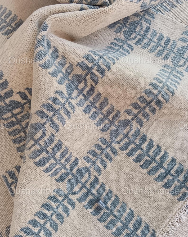 Op de afbeelding: Een close-up van een cr&egrave;mekleurig tapijt met een blauw en wit geometrisch patroon. Het tapijt heeft een franje rand.