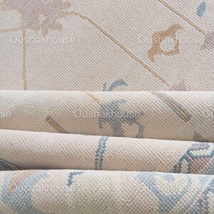 May include: A close-up of a cream-coloured rug with a subtle floral pattern in shades of blue, pink, and beige. The rug is folded in thirds, revealing the texture of the fabric.