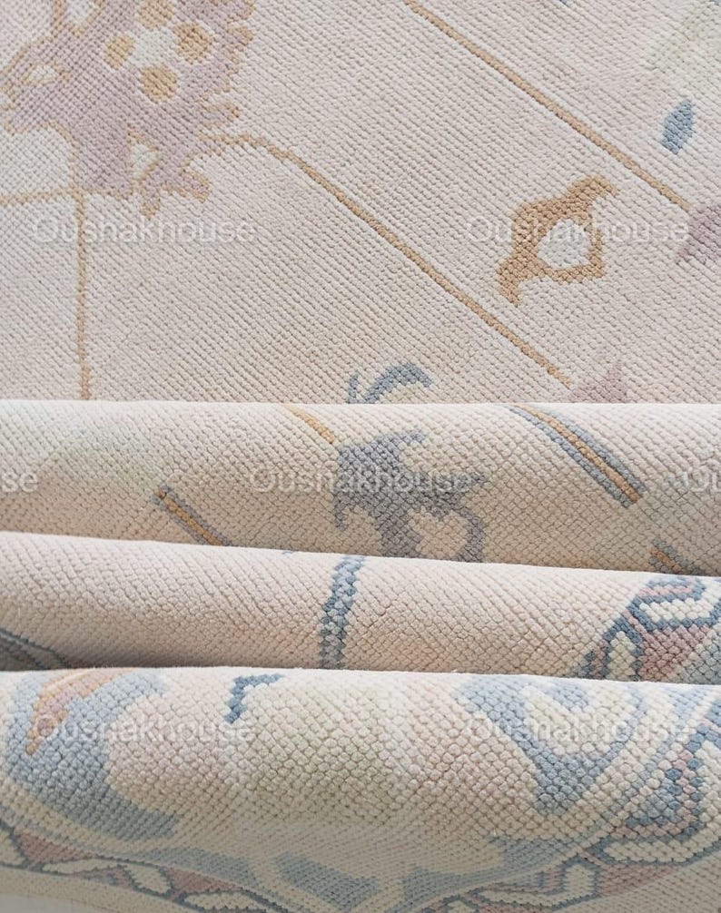 May include: A close-up of a cream-coloured rug with a subtle floral pattern in shades of blue, pink, and beige. The rug is folded in thirds, revealing the texture of the fabric.