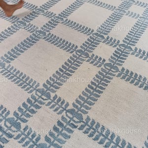 Op de afbeelding: Een wit tapijt met een blauw geometrisch patroon. Het patroon bestaat uit herhalende bladeren.