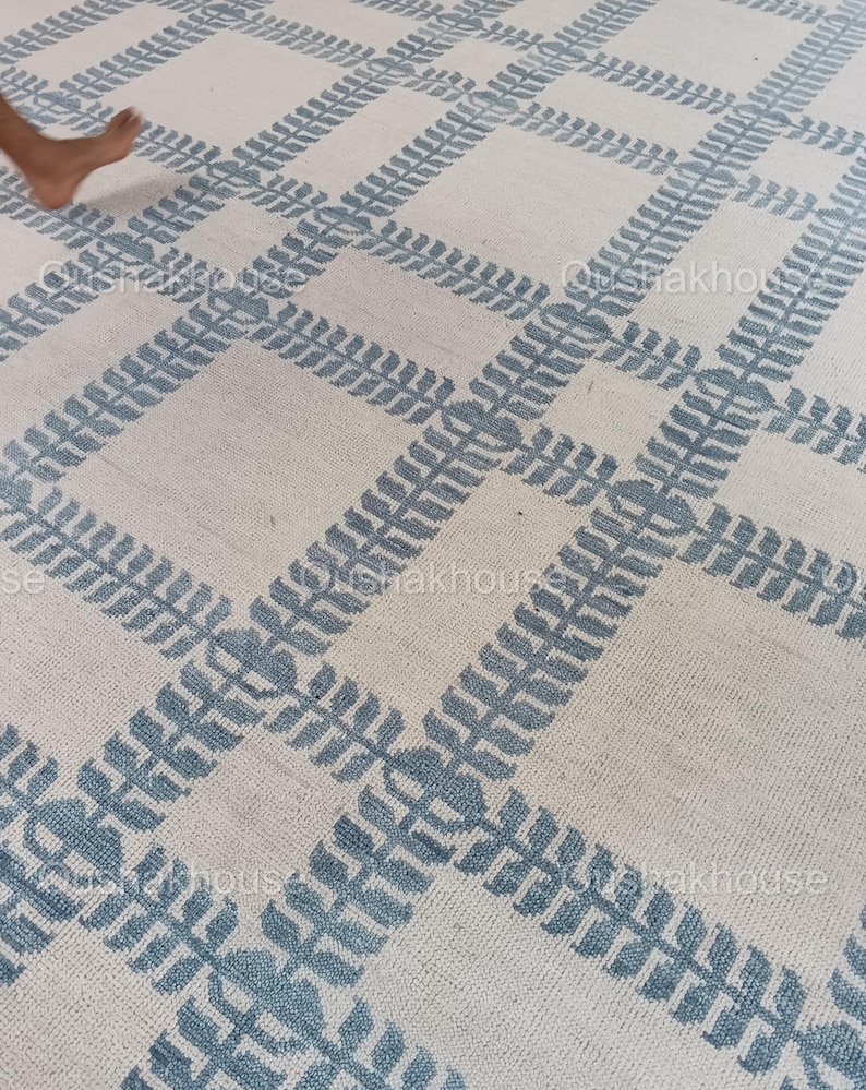 Op de afbeelding: Een wit tapijt met een blauw geometrisch patroon. Het patroon bestaat uit herhalende bladeren.