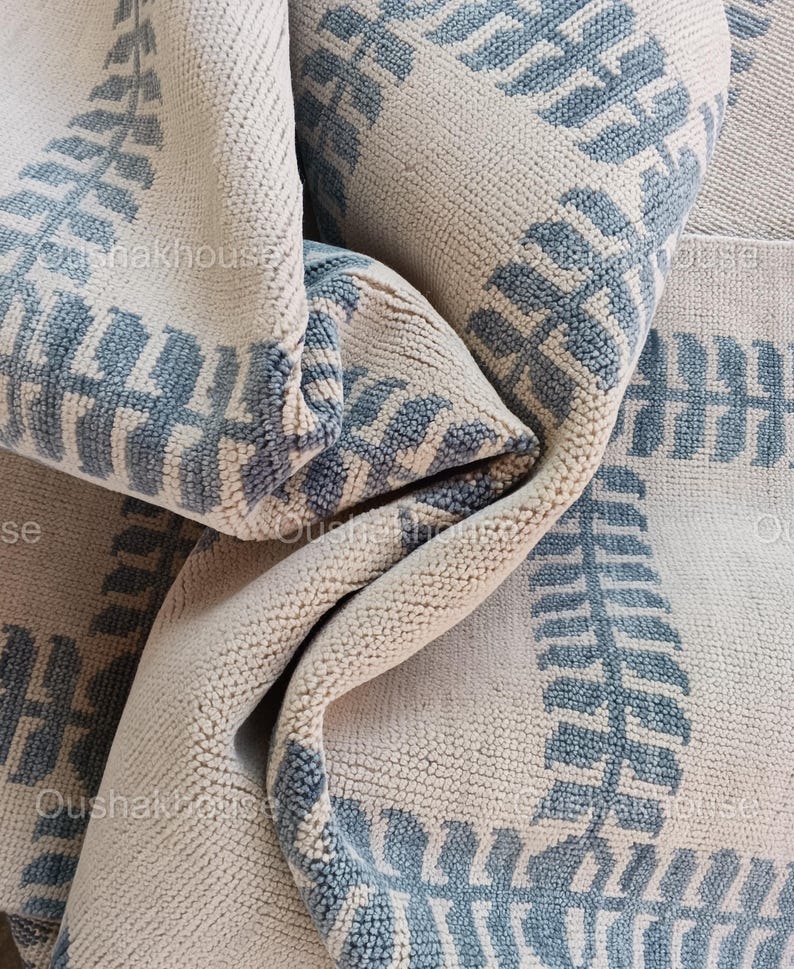 Op de afbeelding: Close-up van een cr&egrave;mekleurig tapijt met een blauw varenbladpatroon. Het tapijt is dubbelgevouwen, waardoor de textuur van de stof zichtbaar is.
