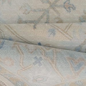 Op de afbeelding: Een opgerolde vloerkleed met een crèmekleurige achtergrond en een vervaagd bloemenpatroon in blauwe en bruine tinten. Het kleed heeft een getextureerd oppervlak en een gefranjerde rand. De afbeelding toont het opgevouwen kleed.