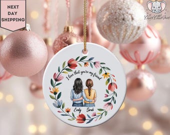 May include: A white ceramic ornament with a floral wreath design and the text "I love that you're my friend." The ornament features two figures embracing. The names "Emily" and "Sarah" are below the figures. Pink and gold Christmas ornaments are in the background.
