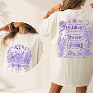 May include: An off-white t-shirt featuring purple graphic prints. The front displays 'OWENS SISTERS APOTHECARY EST 1892' with botanical illustrations. The back features text about salt, rosemary, and lavender with celestial and floral motifs.