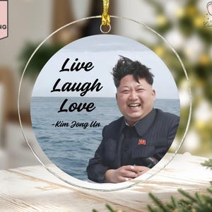 May include: Clear acrylic Christmas ornament with a gold hanging loop. The ornament features a photo of a smiling man with the words "Live Laugh Love - Kim Jong Un" in black text. The background shows a body of water.