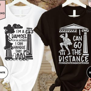 May include: Two t-shirts with a Greek mythology theme. The white t-shirt has a woman standing in front of a column with a dragon and the text "I'm a Damsel I'm in Distress I Can Handle This". The black t-shirt has a man standing in front of a column with the text "I Can Go The Distance".