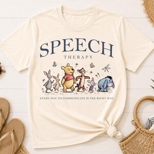 May include: A cream-colored short-sleeve t-shirt featuring the dark blue text SPEECH THERAPY and the phrase EVERY WAY TO COMMUNICATE IS THE RIGHT WAY. The graphic depicts Winnie the Pooh, Tigger, Piglet, Eeyore, and other characters walking.