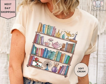 May include: A cream colored t-shirt with a graphic of a bookshelf filled with books and various Disney-themed items, including a teapot, a lamp, a crown, and a flower. The text "Soft Cream" is printed on the bottom right corner of the shirt.