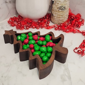 Puede incluir: Un cuenco de madera con forma de árbol de Navidad lleno de caramelos rojos y verdes. Una jarra blanca, una figura de Papá Noel y una guirnalda de cuentas rojas están en el fondo. El cuenco es de madera oscura.
