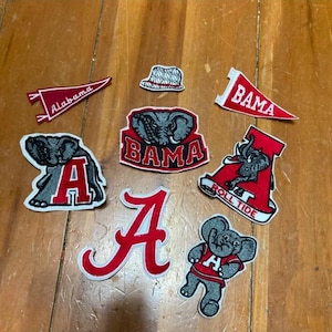 May include: A collection of Alabama Crimson Tide patches and pennants. The patches feature the team's elephant mascot, the letter "A", and the words "Bama" and "Roll Tide" in red and white. A small hat patch is also included.