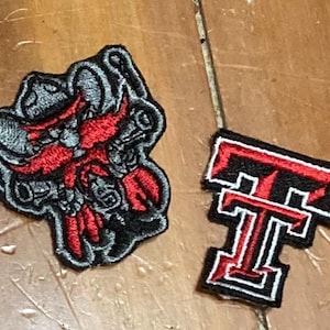 May include: Two embroidered patches. One is a cartoon character with a red beard and a black and gray design. The other is a red and black letter "T" with a white outline.