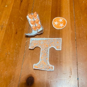 May include: Three embroidered patches: a blue boot with orange butterfly accents, a white star on an orange circle, and a white "T" with an orange floral pattern.