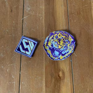 May include: Two embroidered patches, one with a purple and yellow tiger face and the other with a purple and yellow "SW" logo.
