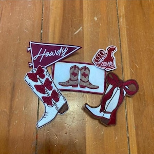 May include: Five embroidered patches with a maroon and white color scheme. The patches include a banner with the word "Howdy", a pair of cowboy boots, a thumbs up with the text "Gig 'em Aggies", and two cowboy boots with bows.