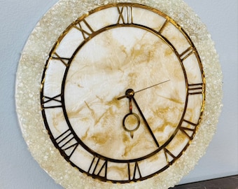 May include: A round, white clock with gold Roman numeral markings and a gold frame. The clock face is textured with a white and gold marbled design. The clock is surrounded by a ring of small, white crystals.