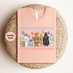 A pink t-shirt with a graphic design featuring various characters from popular media, including a character with a balloon, a character in a white dress, a character in a black suit, and a character in a yellow suit.