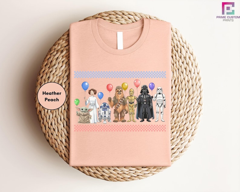 A pink t-shirt with a graphic design featuring various characters from popular media, including a character with a balloon, a character in a white dress, a character in a black suit, and a character in a yellow suit.