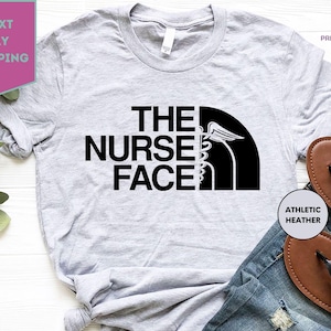 May include: Athletic heather gray t-shirt with the text "THE NURSE FACE" in bold black letters. The design incorporates a medical symbol within a black semi-circle. The shirt is displayed with blue jeans and brown sandals.