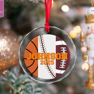 May include: A clear, round Christmas ornament with a red ribbon. The ornament features a basketball, baseball, and football design with the name "JOHNSON" and the year "2025" in orange lettering. The background includes a Christmas tree and a nutcracker.