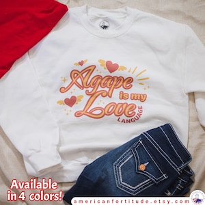 May include: White sweatshirt with a graphic design that says "Agape is my Love Language" in a retro font with hearts and wings. The sweatshirt is available in four colors.