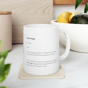 May include: White ceramic mug with a definition of "Rook-Rage" printed on it. The definition reads: "A fit of ragey pouting in response to having one's plans foiled during a game of Rook."  The mug also includes a quote: "Don't mind him, he's just experiencing some rook-rage because he thought all the trumps were gone but he was wrong."