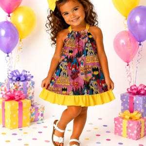 Könnte beinhalten: Ein junges Mädchen trägt ein rosa-gelbes Kleid mit Superhelden-Motiv und den Aufdrucken "Batgirl" und "Wonder Woman". Sie trägt weiße Sandalen und eine gelbe Schleife im Haar. Ballons und Geschenkboxen sind im Hintergrund zu sehen.