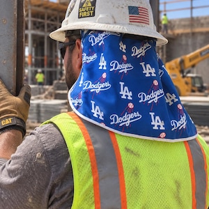 May include: A white hard hat with a Safety First sticker and an American flag decal, featuring a blue neck shade attachment printed with the Los Angeles Dodgers logo, the letters LA, and baseball graphics, worn over a high-visibility yellow vest.