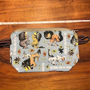 May include: A rectangular, light blue zippered pouch with a brown zipper and handles. The pouch features a repeating pattern of cartoon characters and the text "WHERE THE WILD THINGS ARE". The pouch measures approximately 10 inches in length.