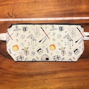 May include: A white pencil case with a black zipper and two white fabric loops. The case is covered in black and red Star Wars themed illustrations, including characters, spaceships, and phrases. The case measures approximately 10 inches in length.